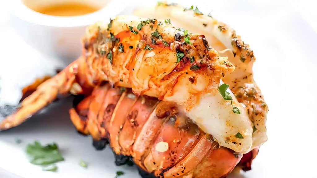 Grilled lobster tail garnished with herbs, served as a festive main dish. Cooked to a golden brown and drizzled with a Christmas-style dipping sauce, it features visible seasoning and fresh parsley sprinkled on top for an exquisite holiday meal.