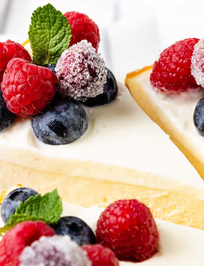 Slices of cheesecake, featured in exquisite dessert recipes, are topped with fresh raspberries, blueberries, and mint leaves. The berries have a sugared coating, complementing the cheesecake's smooth, creamy texture on a crumbly crust. The softly blurred background enhances the presentation.