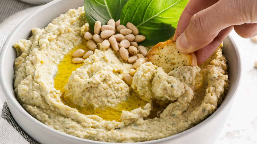 A bowl of hummus, a staple in finger food recipes, is garnished with olive oil, pine nuts, and basil leaves. A hand dips a small piece of bread into the creamy delight.