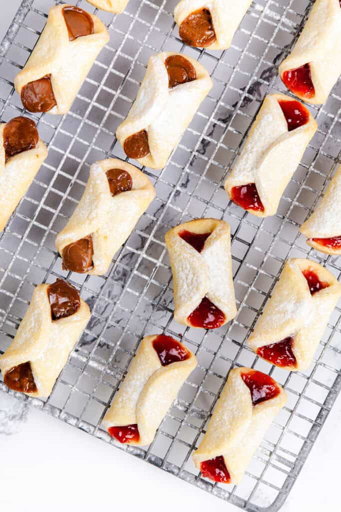 Italian pinch cookies, with their distinctive rectangular shape, are artfully arranged on a wire cooling rack. These treats boast fruit and chocolate fillings, dusted with powdered sugar to enhance their allure. The folded design cleverly reveals a glimpse of the delectable fillings inside.