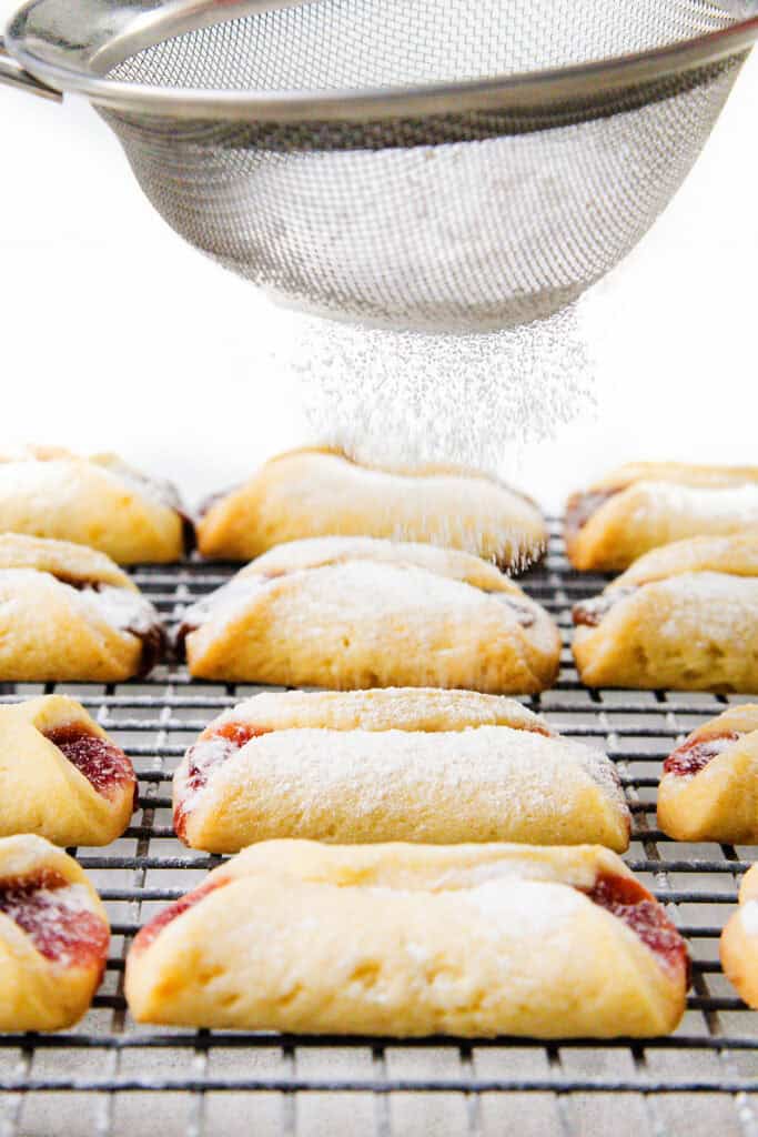 A close-up of Italian pinch cookies cooling on a wire rack. Powdered sugar is being sifted from above, gently dusting the cookies. The golden brown treats reveal red jam peeking out from their pinched edges.