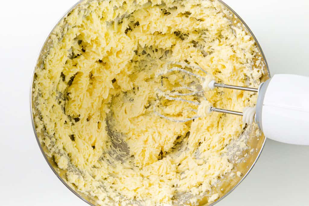 A mixing bowl filled with creamy, light yellow batter for delicious Italian Pinch Cookies. An electric hand mixer with metal beaters is partially visible on the right, showing the batter being mixed. The background is a clean, white surface.