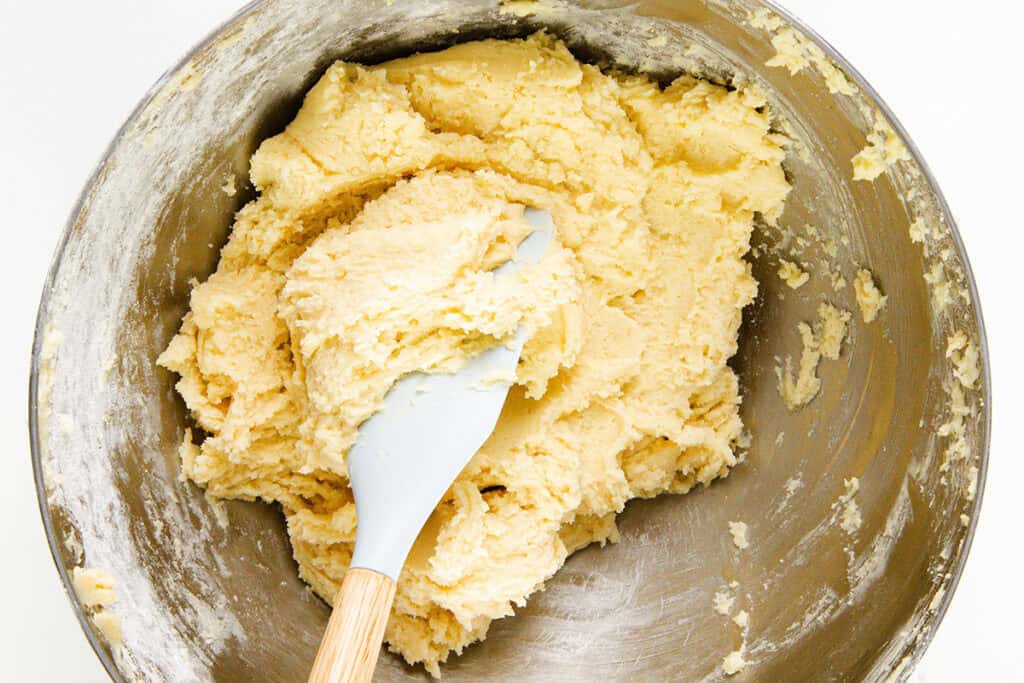 A mixing bowl containing yellow pinch cookie dough with a white spatula partially submerged in it. The Italian-inspired dough has a thick, creamy texture, and the bowl shows traces of flour on its edges. The spatula has a wooden handle.