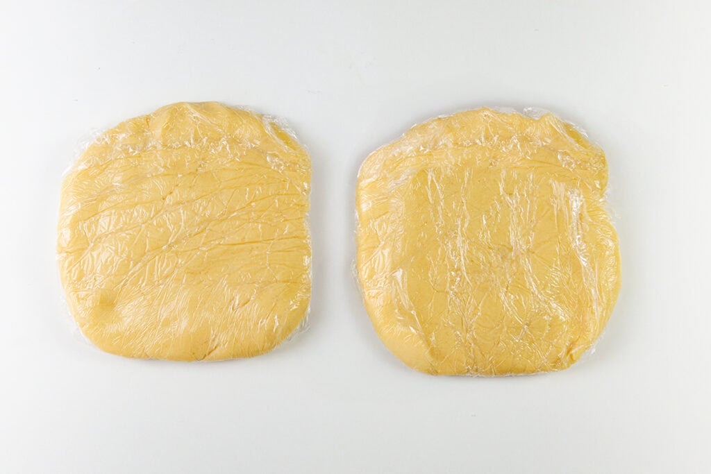 Two round portions of dough, looking as smooth as the finest Italian cookies, are wrapped in clear plastic wrap and placed side by side on a white surface. A gentle pinch reveals their perfectly even shape and impeccable texture.