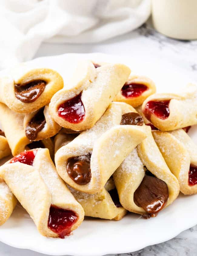 A plate of crescent-shaped Italian pastries, reminiscent of pinch cookies, filled with red and chocolate delights, dusted with powdered sugar. Arranged on a white marble surface, a white cloth and blurred background enhance the cozy atmosphere.