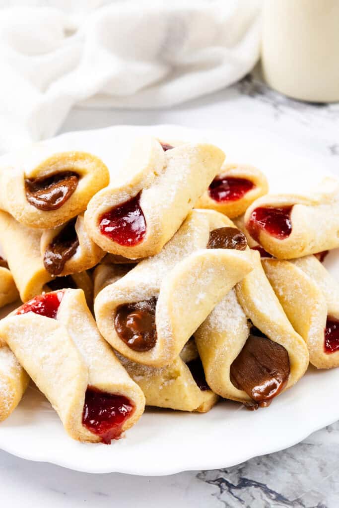 A plate of crescent-shaped Italian pastries, reminiscent of pinch cookies, filled with red and chocolate delights, dusted with powdered sugar. Arranged on a white marble surface, a white cloth and blurred background enhance the cozy atmosphere.