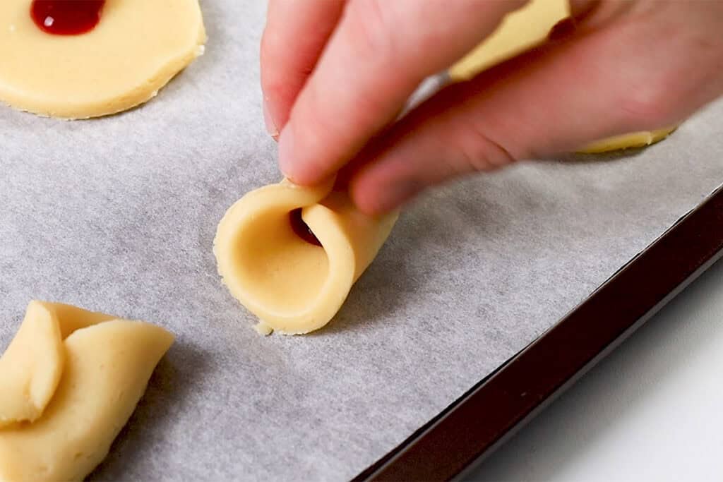 A hand skillfully pinches a piece of dough with red jam inside on a parchment-lined baking sheet, crafting delightful Italian-inspired cookies. Other pieces of dough rest in the background, waiting to be transformed into sweet treats.