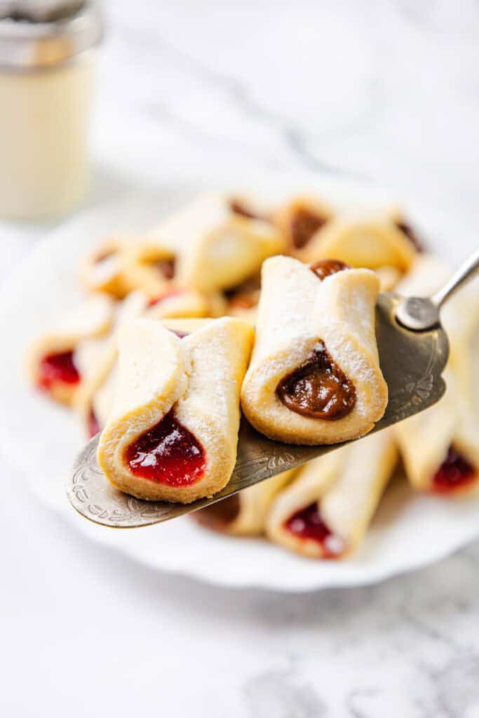 A close-up of pinch cookies filled with red and brown jams on a silver serving utensil. In the background, more delectable pastries are piled high on a white plate, elegantly placed on a marble surface.