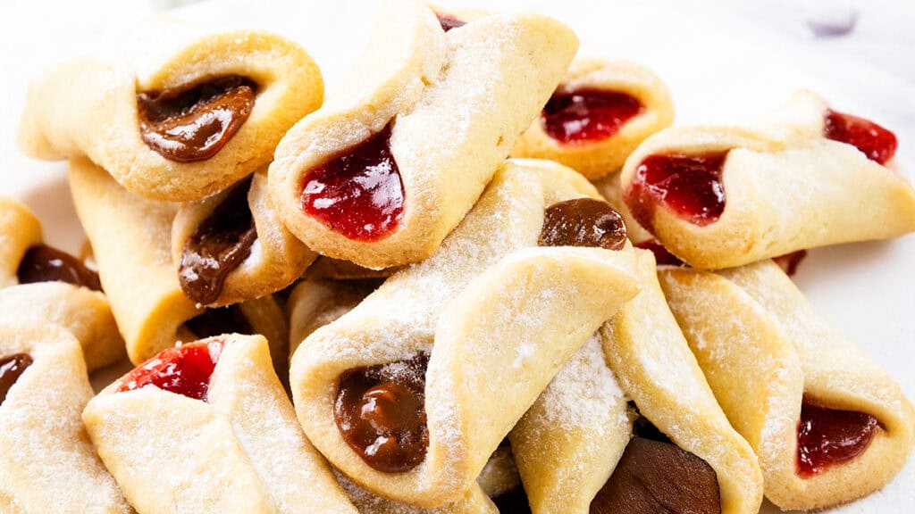 A delightful assortment of Italian pinch cookies, delicately dusted with powdered sugar and filled with rich strawberry and chocolate jams. These pastries boast a golden-brown crust, elegantly presented against a pristine white background.