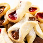 A delightful assortment of Italian pinch cookies, delicately dusted with powdered sugar and filled with rich strawberry and chocolate jams. These pastries boast a golden-brown crust, elegantly presented against a pristine white background.