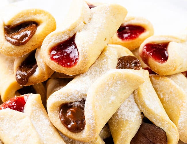 A delightful assortment of Italian pinch cookies, delicately dusted with powdered sugar and filled with rich strawberry and chocolate jams. These pastries boast a golden-brown crust, elegantly presented against a pristine white background.