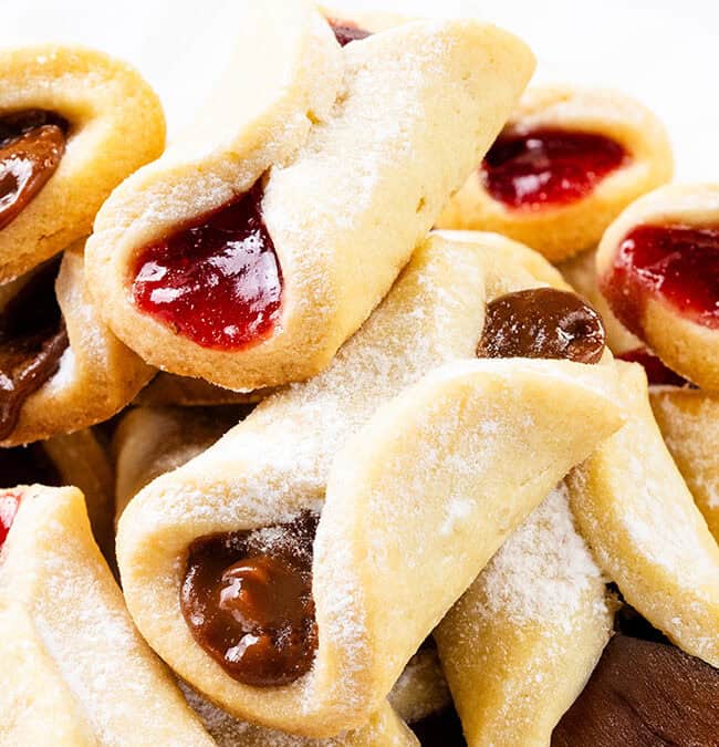 A delightful assortment of Italian pinch cookies, delicately dusted with powdered sugar and filled with rich strawberry and chocolate jams. These pastries boast a golden-brown crust, elegantly presented against a pristine white background.