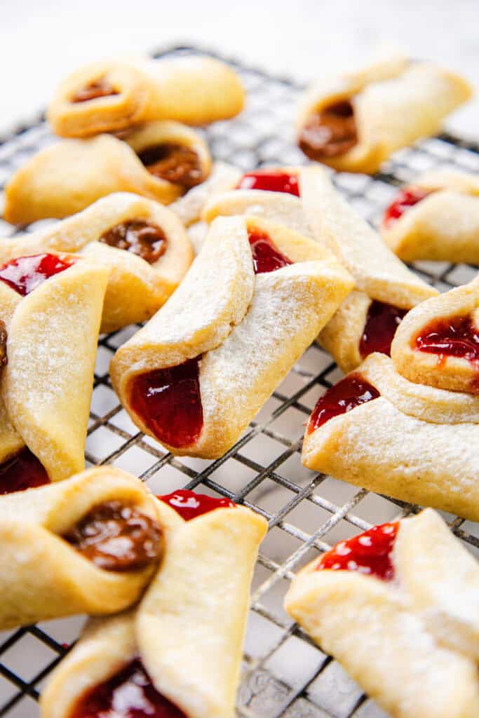 A cooling rack displays an assortment of scrumptious Italian cookies, some brimming with red jam and others with a rich brown filling. These delightful pinch cookies are dusted with powdered sugar and showcase a charming folded design against a blurred white background.