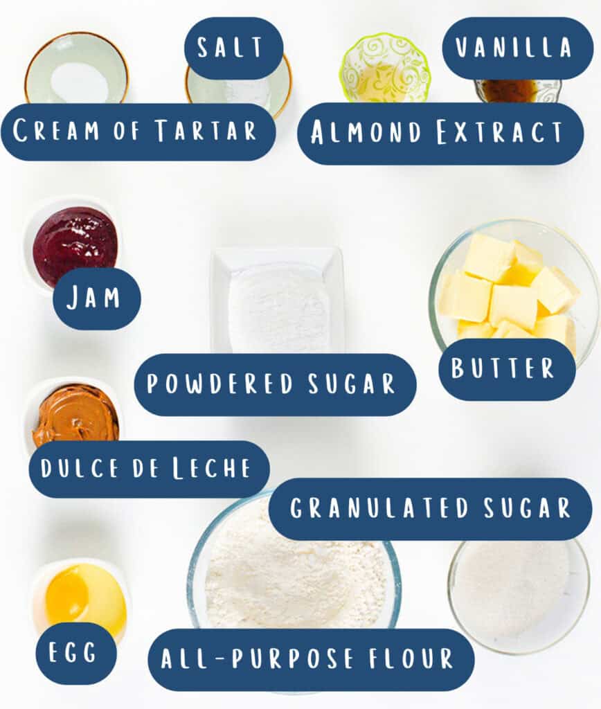 Top view of baking ingredients for Italian Pinch Cookies laid out in bowls, labeled: salt, vanilla, cream of tartar, almond extract, jam, powdered sugar, butter, dulce de leche, granulated sugar, egg, and all-purpose flour on a white background.