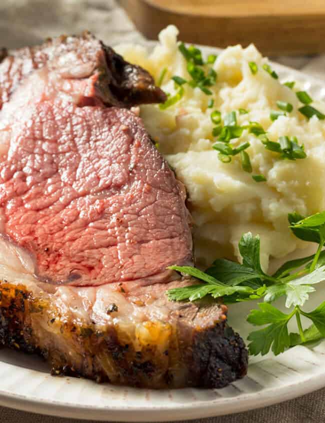 A festive plate of sliced roast beef and creamy mashed potatoes, garnished with chopped green onions and parsley, is perfect for your Christmas table. This delightful addition to your holiday recipes sits invitingly on a light-colored tablecloth.