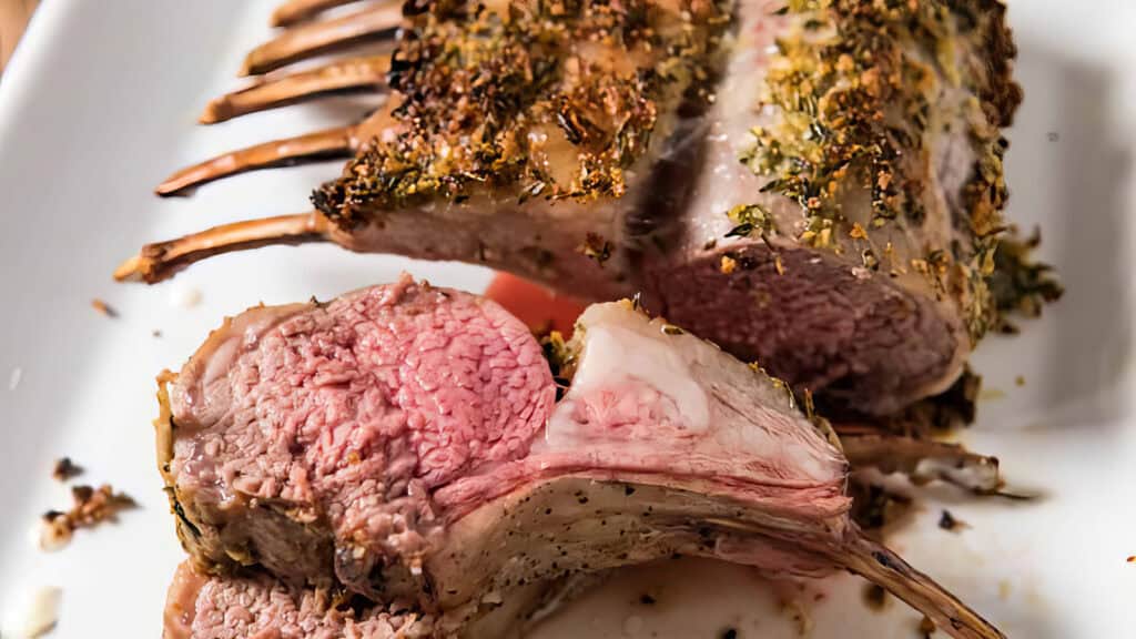 A close-up of a seasoned and cooked rack of lamb on a white plate, perfect for Christmas. One piece is sliced to reveal the juicy, pink interior, with visible herbs and spices crusting this delicious main dish.