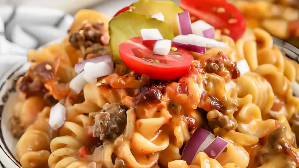 A close-up of a pasta dish featuring curly noodles, ground beef, melted cheese, and garnished with sliced pickles, cherry tomatoes, and diced onions creates a vibrant and appetizing presentation—an enticing addition to any collection of ground beef recipes.