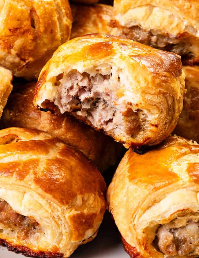 A plate of golden-brown sausage rolls, perfect for Christmas gatherings, some with bites taken out. The flaky pastry is crisp and the filling visibly savory, arranged in a slightly overlapping pile on a white plate—a tempting delight straight from cherished family recipes.