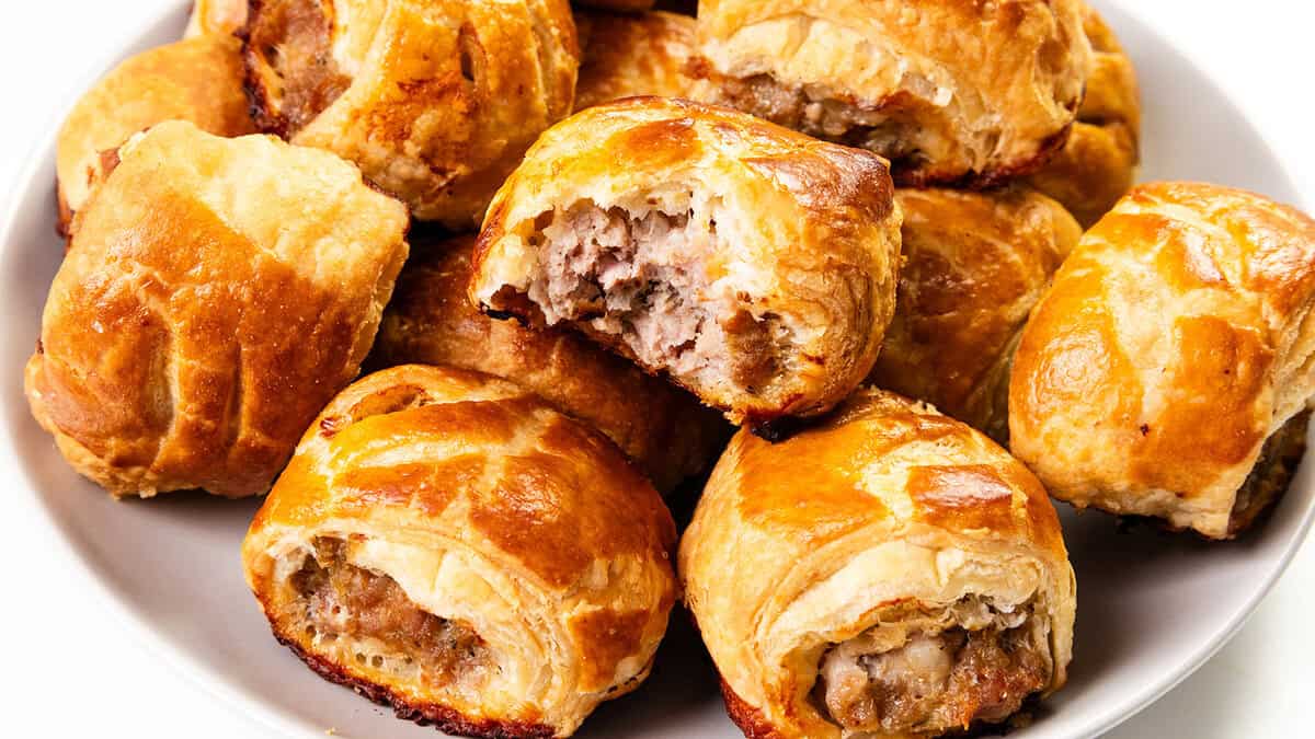 A plate of golden-brown sausage rolls, perfect for Christmas gatherings, some with bites taken out. The flaky pastry is crisp and the filling visibly savory, arranged in a slightly overlapping pile on a white plate—a tempting delight straight from cherished family recipes.