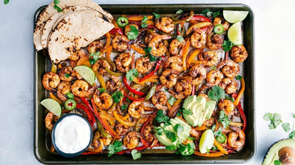 A delicious one pot recipe featuring a sheet pan loaded with seasoned shrimp, sliced red and yellow bell peppers, and onions. It's garnished with lime wedges, jalapeños, cilantro, and avocado slices. Served with grilled tortillas and a small bowl of sour cream on the side.