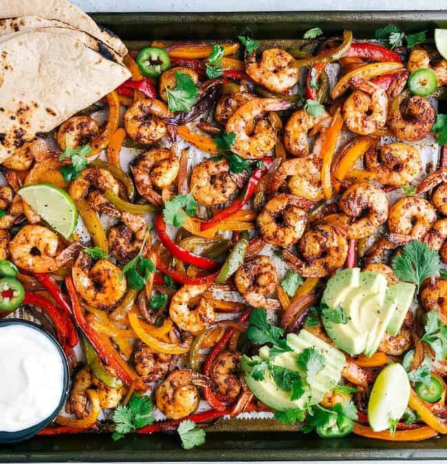 A delicious one pot recipe featuring a sheet pan loaded with seasoned shrimp, sliced red and yellow bell peppers, and onions. It's garnished with lime wedges, jalapeños, cilantro, and avocado slices. Served with grilled tortillas and a small bowl of sour cream on the side.