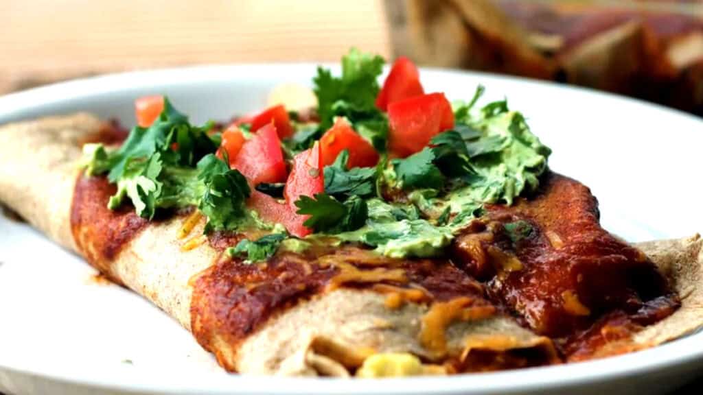 A mouthwatering enchilada topped with melted cheese, chopped tomatoes, guacamole, and fresh cilantro served on a white plate.