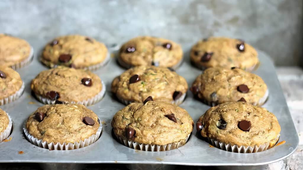 A batch of freshly baked chocolate chip muffins in a baking tray, with some muffins topped with visible chocolate chips. The background is a blurred, textured gray surface.