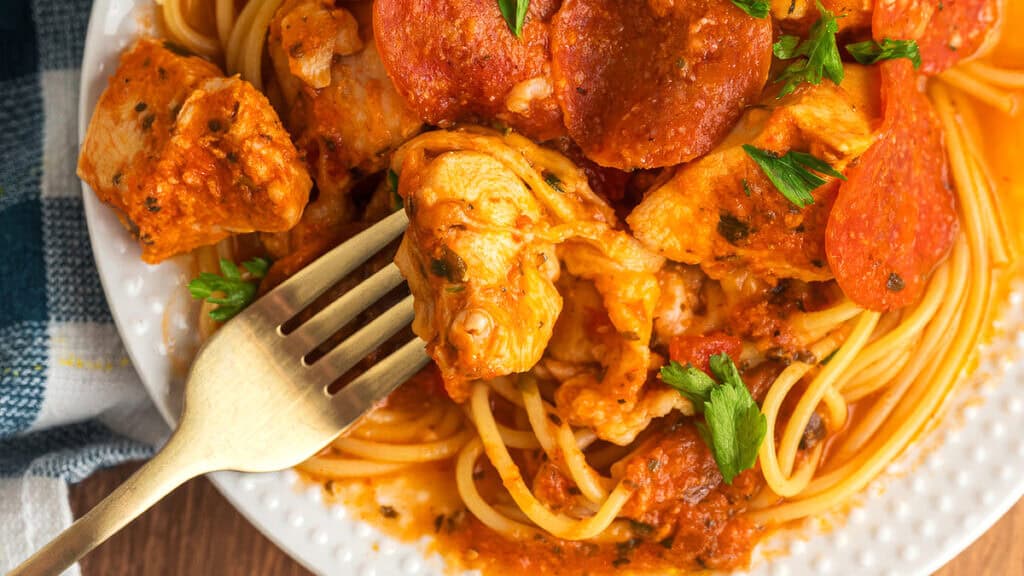 A plate with spaghetti topped with chunks of chicken, pepperoni slices, and tomato sauce. Garnished with parsley, a fork rests beside the food, ready to dig in. A checkered cloth is partially visible at the side.