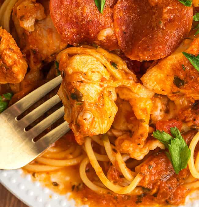 A plate with spaghetti topped with chunks of chicken, pepperoni slices, and tomato sauce. Garnished with parsley, a fork rests beside the food, ready to dig in. A checkered cloth is partially visible at the side.