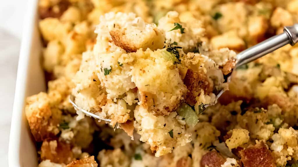 A close-up of a serving of Southern-style stuffing with bread cubes, herbs, and spices being lifted from a casserole dish with a spoon. The dish boasts a golden-brown crust and fluffy texture, embodying the comforting essence of Southern recipes.