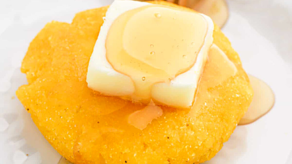 A close-up of a round cornmeal cake, reminiscent of traditional Southern cuisine, topped with a square of butter and drizzled with syrup, set on a white plate.