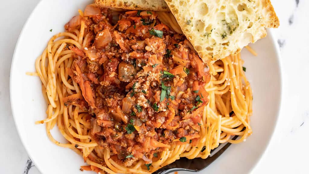 A plate of spaghetti topped with a hearty meat sauce, garnished with chopped herbs. A slice of garlic bread rests on the edge of the plate. The dish is served on a white marble surface.