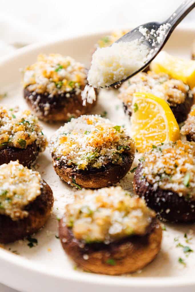 Close-up of stuffed mushrooms adorned with breadcrumbs and herbs. A spoon sprinkles grated cheese over these delightful stuffed appetizers. Lemon wedges sit on the side, while parsley garnishes the white plate, enhancing this savory presentation of Stuffed Mushrooms.