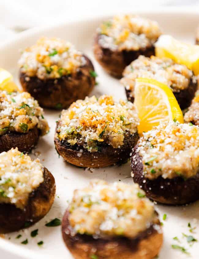 A close-up showcases a plate of stuffed mushrooms, perfectly topped with breadcrumbs and herbs. These delectable stuffed mushrooms are garnished with lemon slices and sprinkled with fresh parsley, all arranged neatly on a pristine white plate.