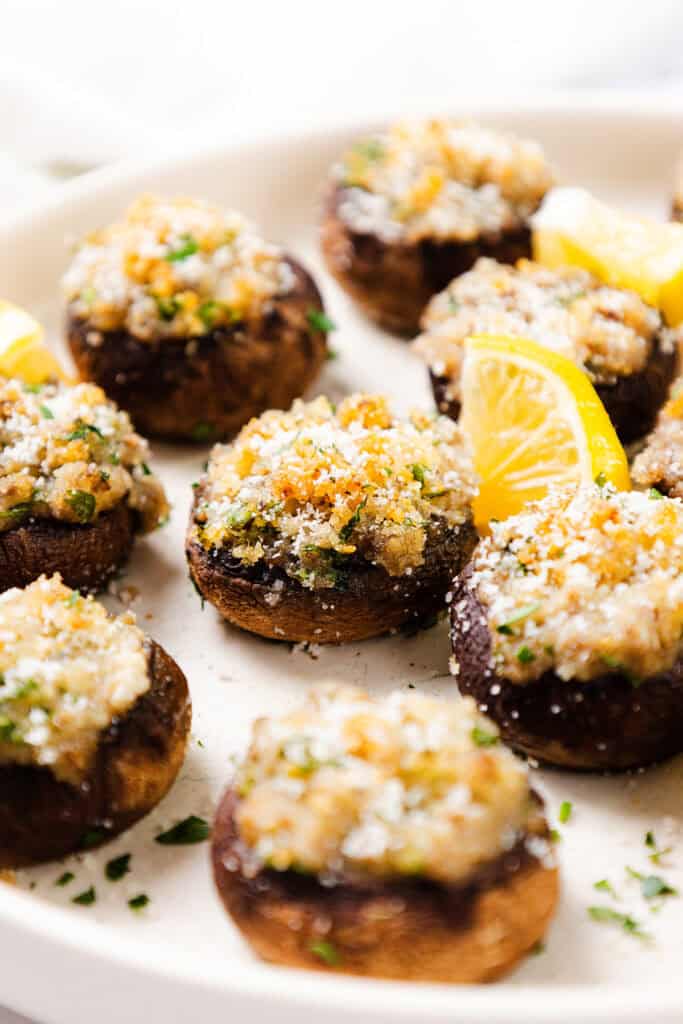 A close-up showcases a plate of stuffed mushrooms, perfectly topped with breadcrumbs and herbs. These delectable stuffed mushrooms are garnished with lemon slices and sprinkled with fresh parsley, all arranged neatly on a pristine white plate.