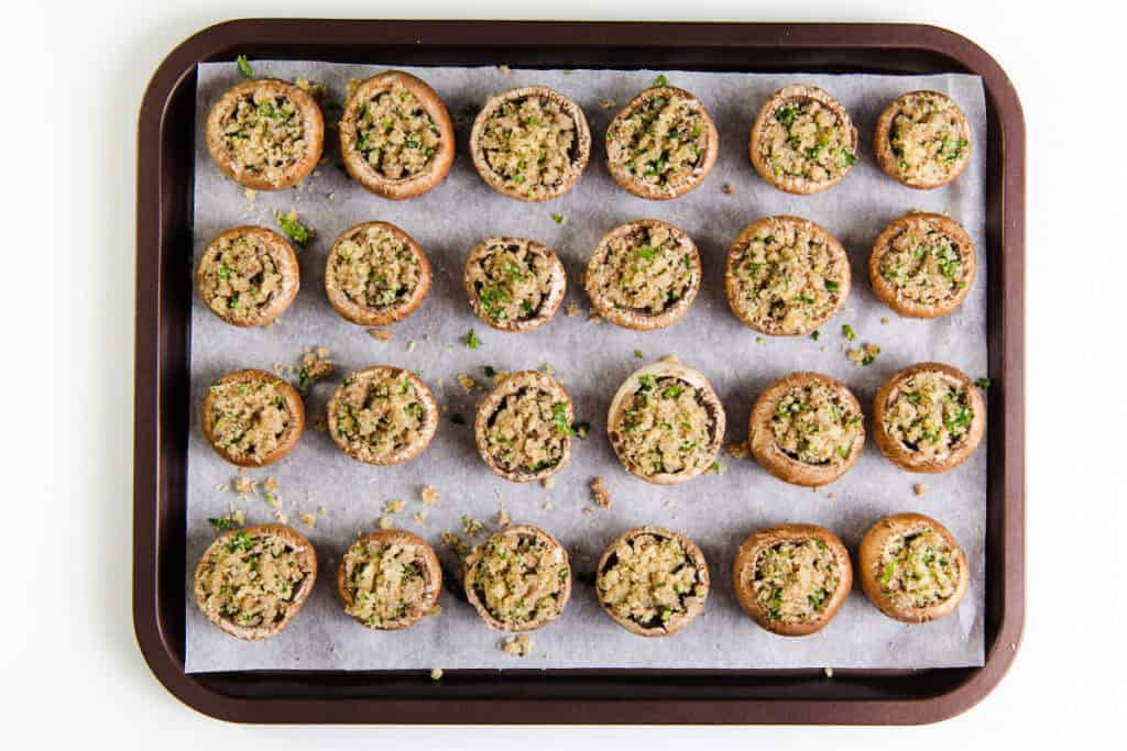 A baking tray lined with parchment paper holds rows of stuffed mushrooms, topped with a mix of breadcrumbs and herbs. This recipe ensures the filling is evenly distributed, and the tray is ready for baking perfection.