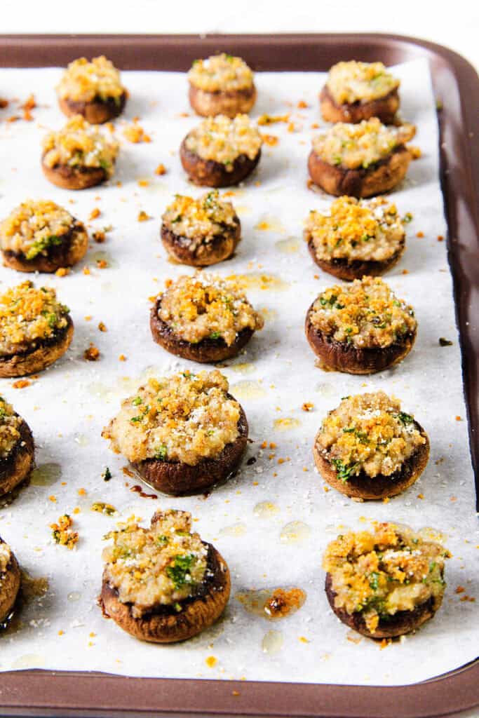 A baking tray lined with parchment paper holds a delicious appetizer of roasted stuffed mushrooms, topped with a golden breadcrumb mixture and fresh herbs. This irresistible recipe features mushrooms drizzled with oil, evenly spaced and lightly browned to perfection.