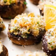 Close-up of stuffed mushrooms artfully arranged, their savory filling complemented by crispy breadcrumbs and herbs. Garnished with grated cheese and lemon wedges, these stuffed mushrooms lie on a pristine white surface, highlighting their delectable toppings.