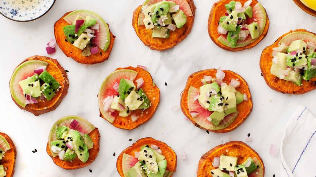 Colorful finger food recipes on a marble surface feature sweet potato slices topped with diced avocado, red onion, and black sesame seeds. The toppings add green and pink hues, creating a vibrant and fresh presentation.