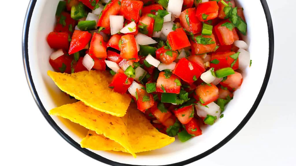 A bowl of fresh salsa containing diced tomatoes, onions, cilantro, and jalapeños. Three yellow tortilla chips rest on the side of the bowl. The dish is placed on a white surface.