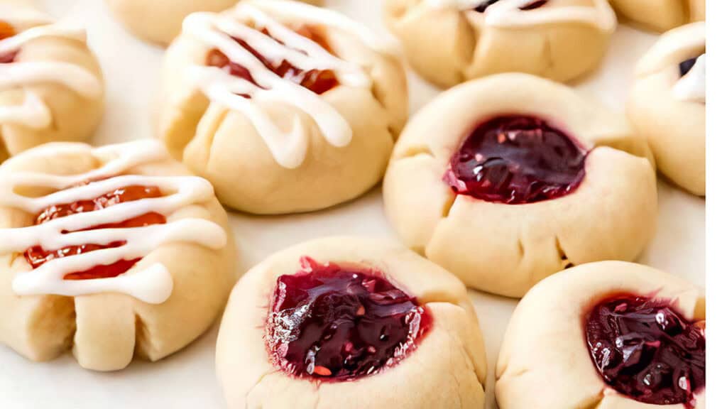 A close-up of freshly baked thumbprint cookies filled with red jam. Some thumb print cookies are drizzled with white icing, creating a variation in texture and color. The cookies are arranged on a white surface.