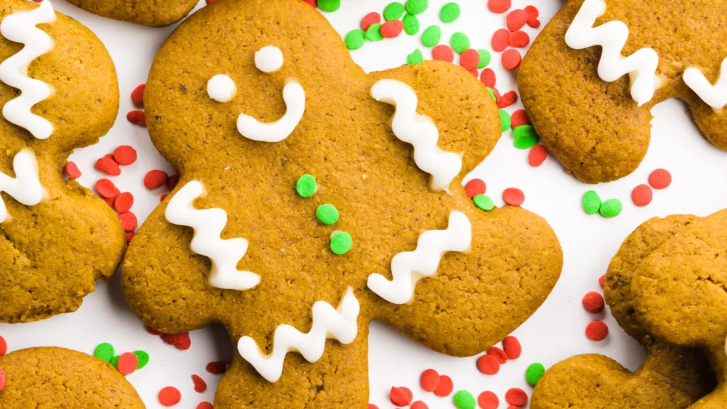 A gingerbread cookie shaped like a person is festively decorated with white icing and green buttons, surrounded by red and green sprinkles on a white background, making it a perfect holiday treat.