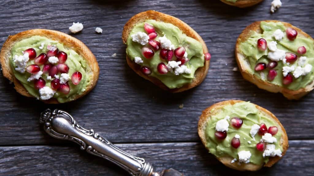 Toasted bread slices topped with creamy avocado, crumbled cheese, and vibrant pomegranate seeds sit invitingly on a dark wooden surface. Perfect as finger food recipes, a vintage silver butter knife rests elegantly beside the arrangement.