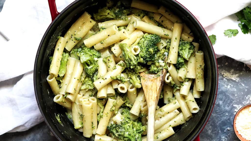 A pot filled with ziti pasta mixed with creamy broccoli sauce, perfect for those seeking skinny recipes. A wooden spoon rests in the pot, surrounded by a white cloth and a small bowl with additional ingredients on a gray surface.