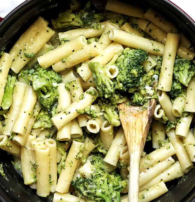 A pot filled with ziti pasta mixed with creamy broccoli sauce, perfect for those seeking skinny recipes. A wooden spoon rests in the pot, surrounded by a white cloth and a small bowl with additional ingredients on a gray surface.