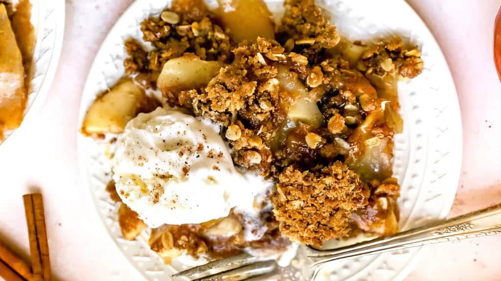 A plate of apple crisp topped with a scoop of vanilla ice cream. The crisp features a crumbly oat topping and is garnished with a sprinkle of cinnamon. A fork rests on the plate's edge.