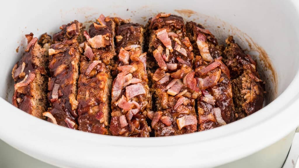 A crockpot filled with sliced meatloaf, crafted from ground beef recipes, is topped with a rich glaze and pieces of bacon. The dish appears juicy and savory, ready to be served.