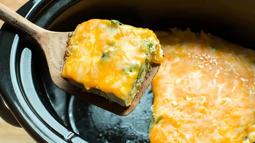 A close-up of a cheesy casserole recipe being lifted from a black slow cooker with a wooden spatula. The casserole is topped with melted cheese, appearing creamy and golden.