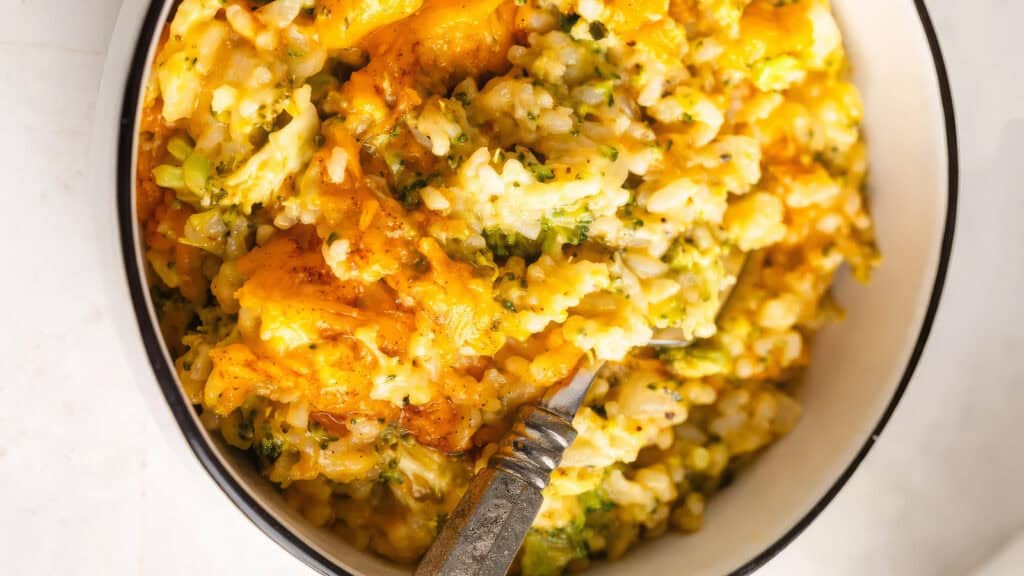 A bowl of creamy baked rice infused with Southern cuisine flair, featuring melted cheddar cheese, broccoli, and visible herbs. A fork is partially embedded in the rich, golden dish, beckoning a taste of this traditional Southern cooking delight.