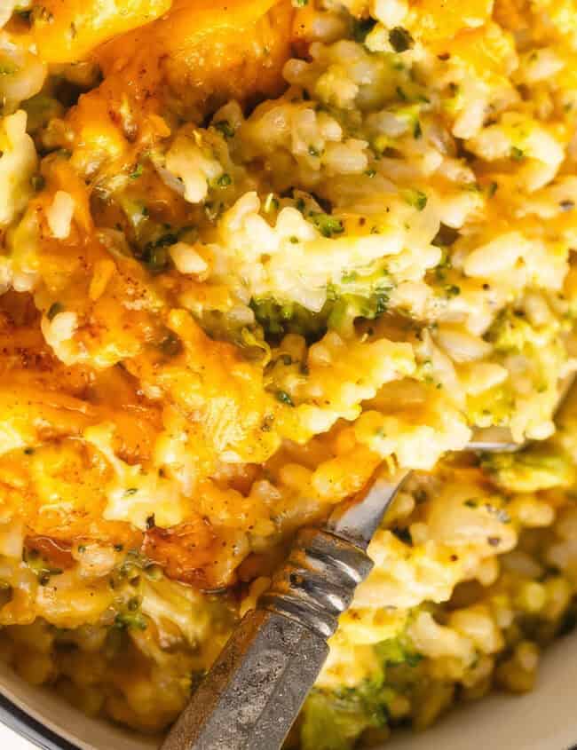 A bowl of creamy baked rice infused with Southern cuisine flair, featuring melted cheddar cheese, broccoli, and visible herbs. A fork is partially embedded in the rich, golden dish, beckoning a taste of this traditional Southern cooking delight.